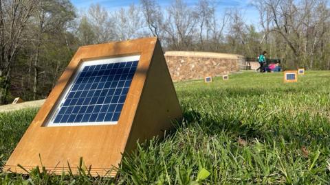 A solar-powered object in a wooden triangular frame sits on vibrant green grass. In the background, bare trees and a stone wall are visible under a clear blue sky.